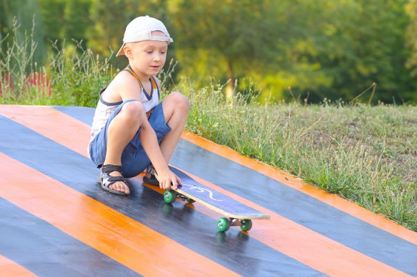 آموزش اسکیت برد کودک پسر ورزش تابستانی در فضای باز با طبیعت در پس زمینه