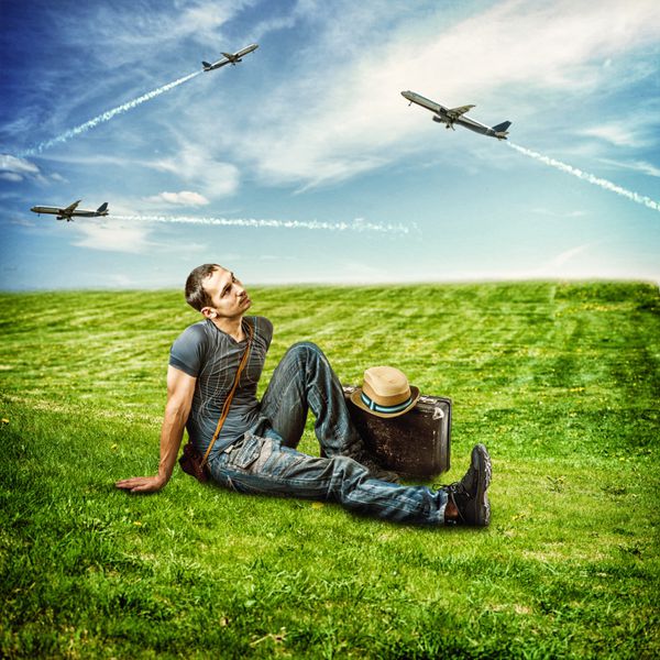 مرد جوان مسافری که روی یک چمن سبز تازه نشسته و به هواپیمای در حال پرواز در آسمان آبی نگاه می کند