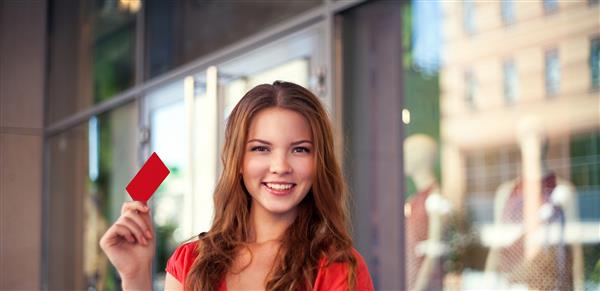 دختر زیبای جوانی که یک کارت اعتباری خالی در فضای باز در مقابل پس زمینه فروشگاه در دست دارد