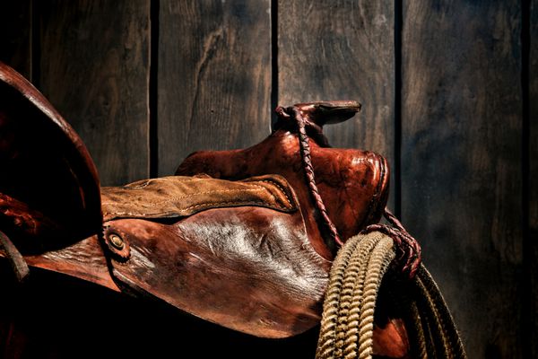 افسانه غول‌پیکر آمریکایی رودئو کابوی طناب‌زنی لاریات کمند آویزان به یک زین وسترن چرم قهوه‌ای اصیل استفاده شده و فرسوده در مزرعه‌داری قدیمی
