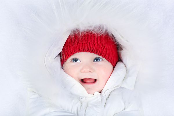 کودک خندان زیبا با چشمان آبی زیبا در کت و شلوار برفی سفید و کلاه بافتنی قرمز گرم