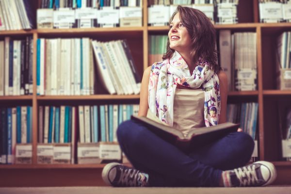 دانش آموز دختر متفکری که کنار قفسه کتاب نشسته و کتابی روی کف کتابخانه دارد