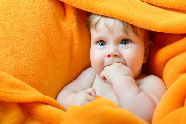 پرتره نوزاد پسر بامزه ای که روی چهارخانه نارنجی دراز کشیده و انگشتش را می مکد
