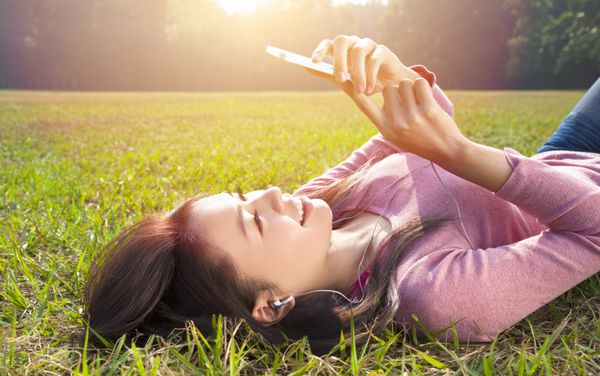 زن جوان خندان در حال لمس تلفن همراه و دراز کشیدن روی چمنزار