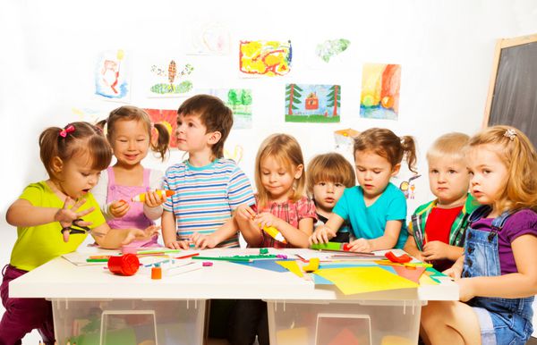گروه بزرگی از بچه های کوچک در حال نقاشی با مداد و چسب زدن با چسب در کلاس هنر در مهد کودک