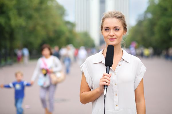 خبرنگار جوان با میکروفون در دست در خیابان ایستاده و به دور نگاه می کند