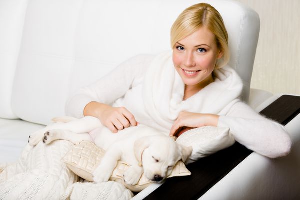 زنی نشسته روی مبل چرمی سفید با خوابیدن روی بالش توله سگ سفید