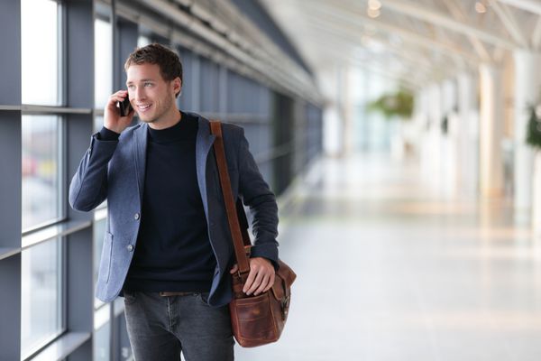 مرد تجاری شهری با تلفن هوشمند صحبت می کند و در حال قدم زدن در داخل فرودگاه است تاجر جوان با کت و شلوار و کیف شانه ای مدل مرد خوش تیپ در 20 سالگی