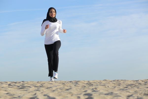 نمای جلوی یک زن اماراتی عربی در حال دویدن در ساحل با افق در پس زمینه