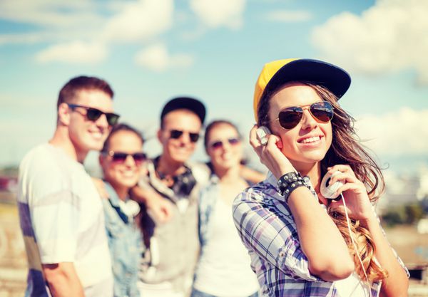 تعطیلات تابستانی و مفهوم نوجوانی - دختر نوجوان با عینک آفتابی کلاه و هدفون در حال معاشرت با دوستان در بیرون