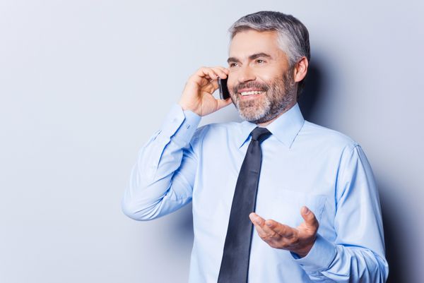 به اشتراک گذاری خبرهای خوب مرد بالغ شاد با پیراهن و کراوات در حال صحبت با تلفن همراه و ژست گرفتن در حالی که در پس زمینه خاکستری ایستاده است