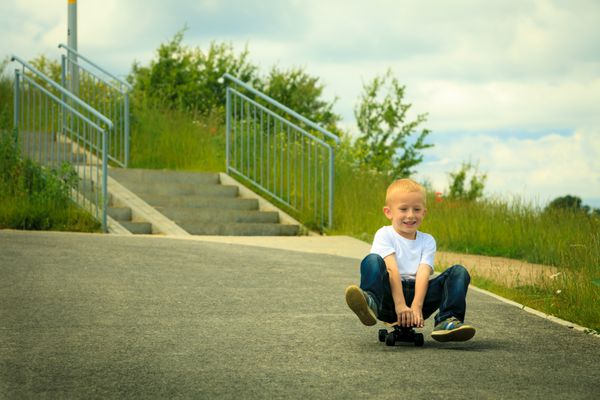 کودکی فعال مرد کوچک اسکیت بورد کودک پسر اسکیت باز در کوچه پارک روی اسکیت بورد می نشیند در فضای باز