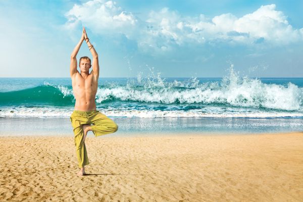 مرد جوانی در حال انجام یوگا و مدیتیشن در موقعیت درخت در ساحل دریا