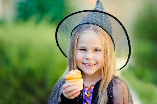 دختر کوچک شادی که لباس جادوگر پوشیده است در جشن هالووین کیک کوچک می خورد