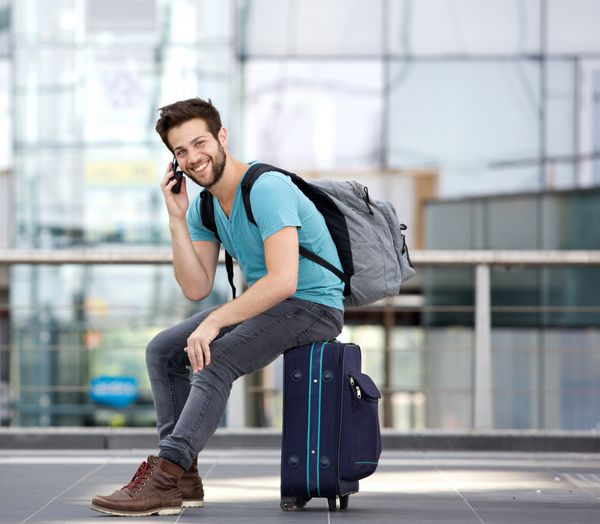 پرتره یک مرد جوان خوش تیپ که روی چمدان نشسته و با تلفن همراه در فرودگاه تماس می گیرد