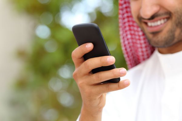دست مرد عرب سعودی خندان با استفاده از تلفن هوشمند در فضای باز در پارکی با پس زمینه سبز