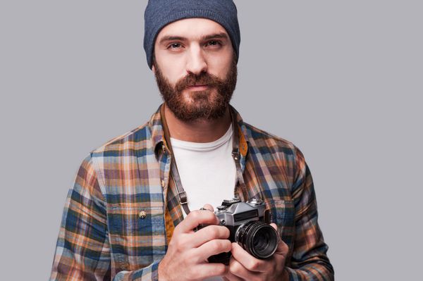 عکاس با اعتماد به نفس مرد جوان ریشو و خوشتیپی که دوربین قدیمی را در دست گرفته و در حالی که در مقابل پس زمینه خاکستری ایستاده به دوربین نگاه می کند