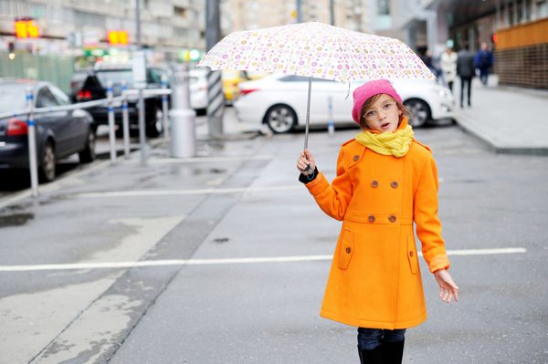 دختر بچه مدرسه ای شایان ستایش و شیک با کت نارنجی روسری زرد و کلاه صورتی و چکمه هایی با چتر رنگارنگ در حال قدم زدن در خیابان شهر در روز پاییزی