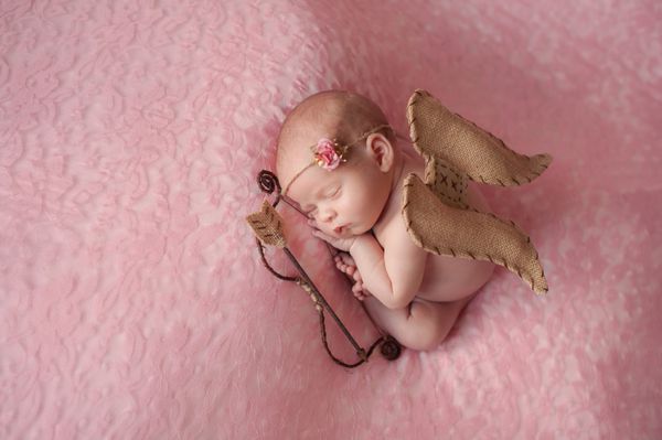 پرتره دختر تازه متولد شده ده روزه او لباس کوپید با بال‌های فرشته کمان و تیر به تن دارد و روی مواد صورتی روشن خوابیده است