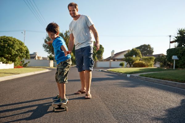 پسر جوانی که در حال یادگیری اسکیت‌برد سواری است در حالی که پدر به او در خیابان حومه شهر می‌آموزد و سرگرم می‌شود