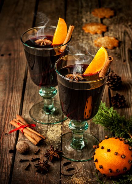 در یک لیوان با تکه های پرتقال بادیان و چوب دارچین روی میز چوبی قدیمی قرار داده شده است نوشیدنی گرم کننده کریسمس یا زمستان با مواد اولیه دستور پخت در اطراف