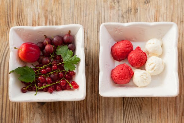 توت‌های قرمز تازه و توپ‌های کوچک کاسه‌های مربعی سفید بستنی خوشمزه روی یک میز چوبی قدیمی