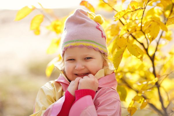 پرتره ای نزدیک از یک دختر کوچک زیبا با کت و کلاه صورتی روشن در میان برگ های زرد در یک روز آفتابی پاییزی