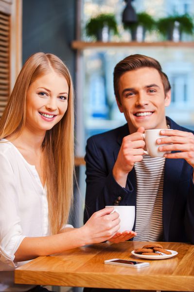 ما عاشق گذراندن وقت با هم هستیم زوج جوان زیبا که به دوربین نگاه می کنند و لبخند می زنند در حالی که در کافه با هم از قهوه لذت می برند