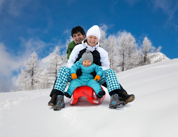 خانواده جوان خندان و شاد سه نفره سورتمه سواری تفریح در کوه های آلپ پیست اسکی در حال حرکت