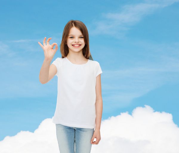 تبلیغات رویا ژست کودکی و مردم - دختر کوچک خندان با تی شرت سفید سفید که علامت ok را روی پس زمینه آسمان آبی نشان می دهد