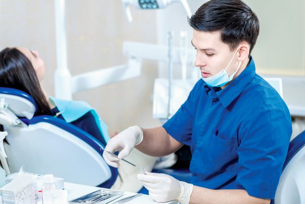 دندانپزشک در محل کار دکتر دندانپزشک با اعتماد به نفس ابزارهایی را برای درمان بیمار در حالی که بیمار روی صندلی دندانپزشکی نشسته است انتخاب می کند