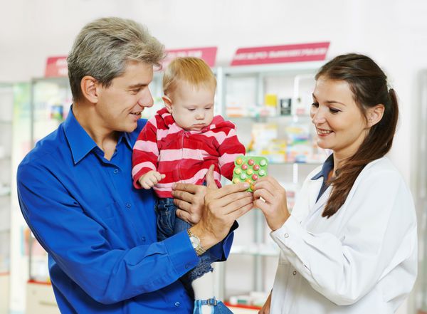 شیمیدان داروساز زن شاد و بانشاط در حال نشان دادن قرص ویتامین به پدر مرد با نوزاد دختر کوچک در داروخانه داروخانه