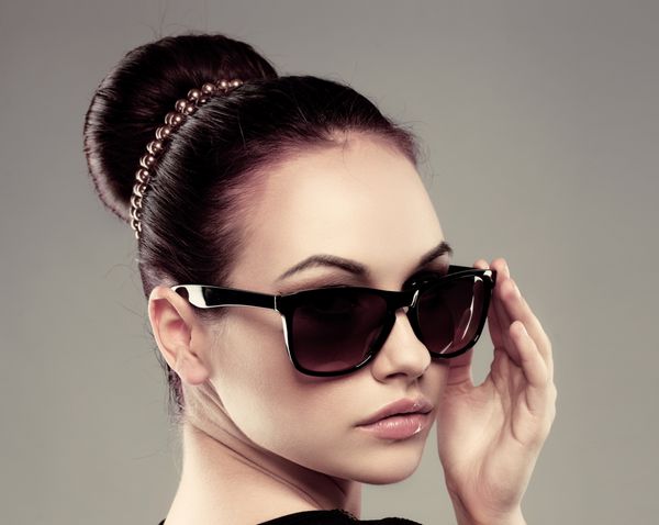نمای نزدیک از مدل زیبا در عینک آفتابی مشکی شیک زن جوان زیبا با مدل موی رترو که در استودیو ژست گرفته است