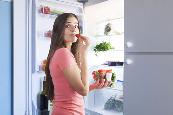 زن جوان زیبا در حال خوردن توت فرنگی از یخچال مفهوم تغذیه سالم