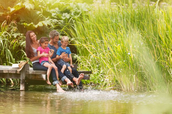 تابستان پرتره یک خانواده شاد که روی لبه یک پانتون چوبی نشسته اند پاهایشان در رودخانه است و آب می پاشند