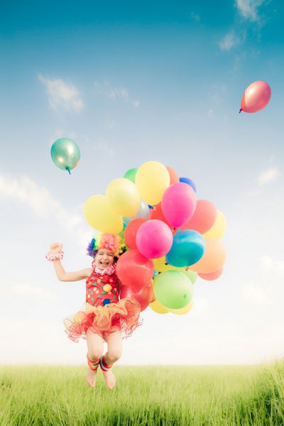کودک شاد در حال پریدن با بادکنک های اسباب بازی رنگارنگ در فضای باز بچه خندان در حال تفریح در زمین سبز بهار در برابر پس زمینه آسمان آبی مفهوم آزادی