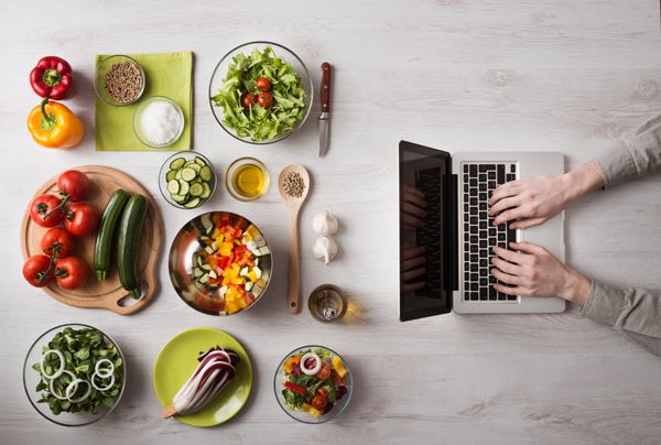 مردی در آشپزخانه در حال جستجوی دستور العمل هایی در لپ تاپ خود با مواد غذایی و سبزیجات تازه در سمت چپ نمای بالا