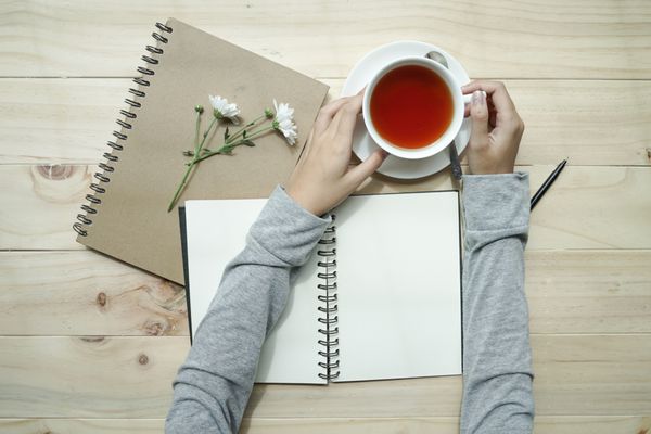 دست های زن که فنجان چای را در دست گرفته و دفترچه خالی روی میز چوبی باز شده است نمای بالا مفهوم نوشتن