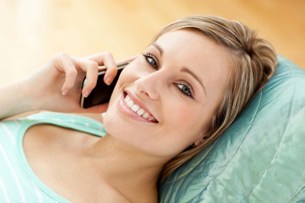 زن جوان شادی که روی مبل خانه دراز کشیده با تلفن صحبت می کند