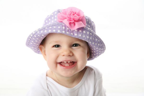 دختر بچه خندان در حال نشان دادن دندان هایی با کلاه خالخالی بنفش با گل صورتی جدا شده روی پس زمینه سفید