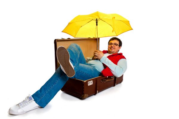 مرد جوانی با عینک و جلیقه قرمز در یک صندوق عقب بزرگ عتیقه در استودیو روی پس‌زمینه سفید دراز کشیده است