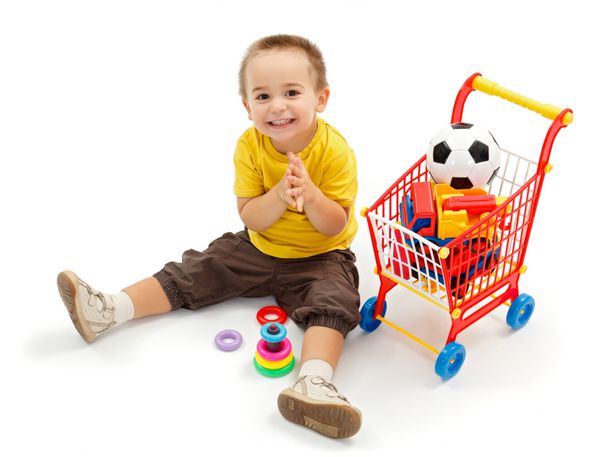 پسر کوچولوی شادی که روی زمین نشسته و اسباب بازی های جدید در سبد خرید کوچک