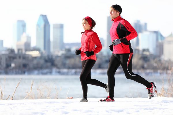 دونده هایی که در برف زمستانی با پس زمینه خط افق شهر می دوند زوج جوان چند نژادی سالم دونده زن آسیایی و مرد قفقازی در حال دویدن با خط افق مونترال کبک کانادا