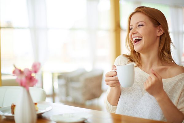 تصویر زن جوان و زیبا با فنجان قهوه در حال خندیدن در کافه