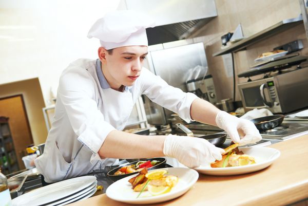 سرآشپز مرد جوان با لباس فرم سفید که غذا را روی بشقاب در آشپزخانه تجاری رستوران تزئین می کند