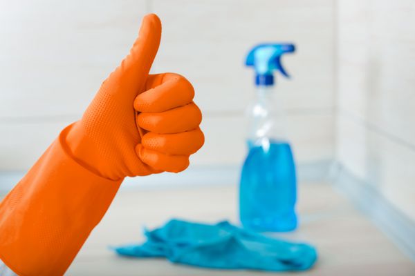 نمای نزدیک از دست انسان با دستکش لاستیکی نارنجی که علامت ok را در برابر بطری اسپری نشان می دهد و دستشویی روی میز آشپزخانه تمیز