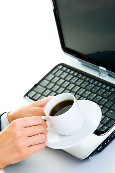 نمای نزدیک از دست‌های تاجر زن که یک فنجان قهوه را روی لپ‌تاپ باز گرفته است