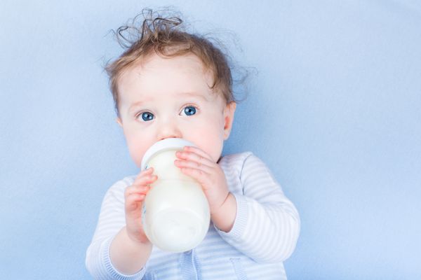 نوزاد کوچولوی شیرین با چشمان آبی زیبا در حال نوشیدن شیر در یک بطری پلاستیکی در حال استراحت روی یک پتوی بافتنی آبی