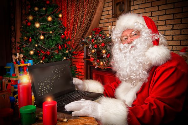 بابا نوئل با لپ تاپ در خانه اش پشت میز نشسته است زمان کریسمس