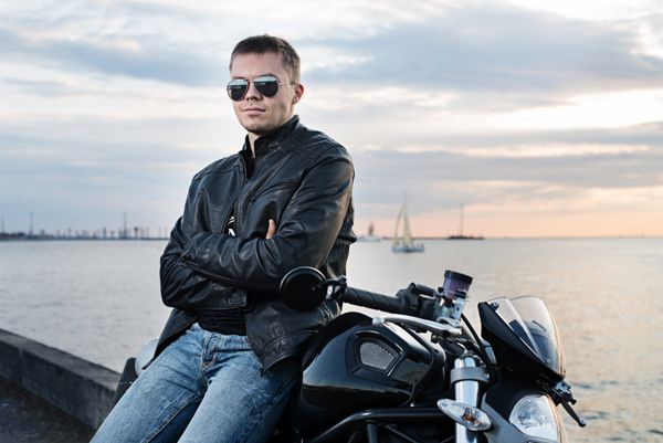 مرد جوان خوشتیپ با ژاکت چرمی سوار بر موتور سیکلت در نور غروب آفتاب بر روی خاکریز دریا